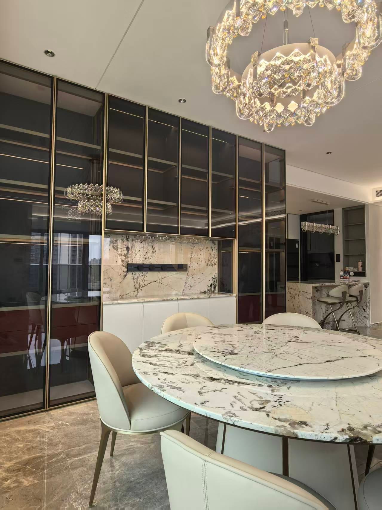 Round marble dining table under a crystal chandelier before a bronze-framed glass display wall