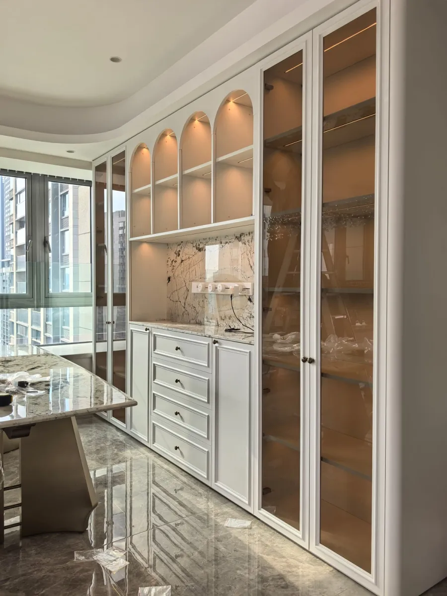 Arched dining display wall with lit glass columns, marble slab niche and white paneled stainless steel cabinetry