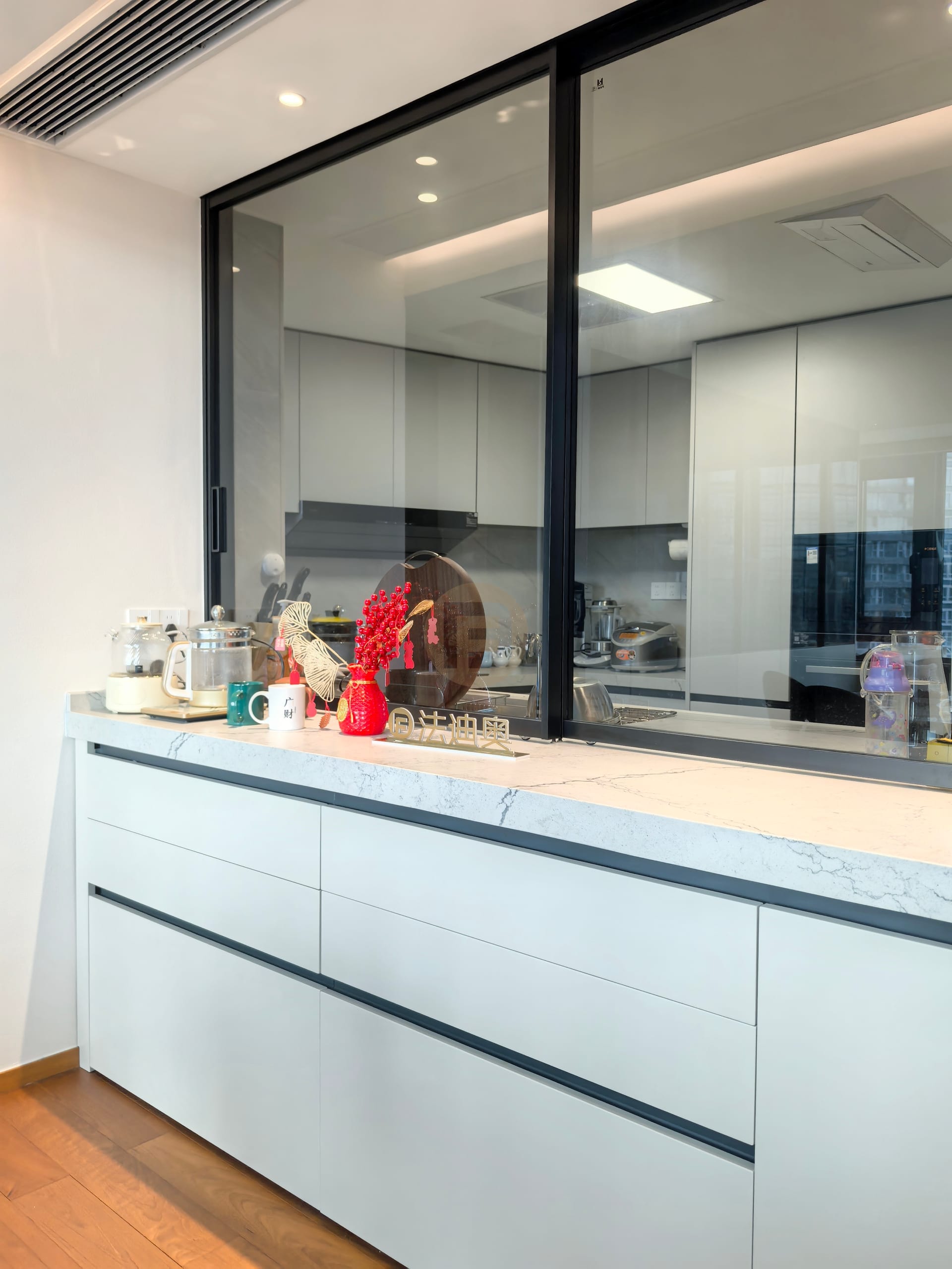 Stainless steel servery cabinetry with marble worktop below a black-framed pass-through window