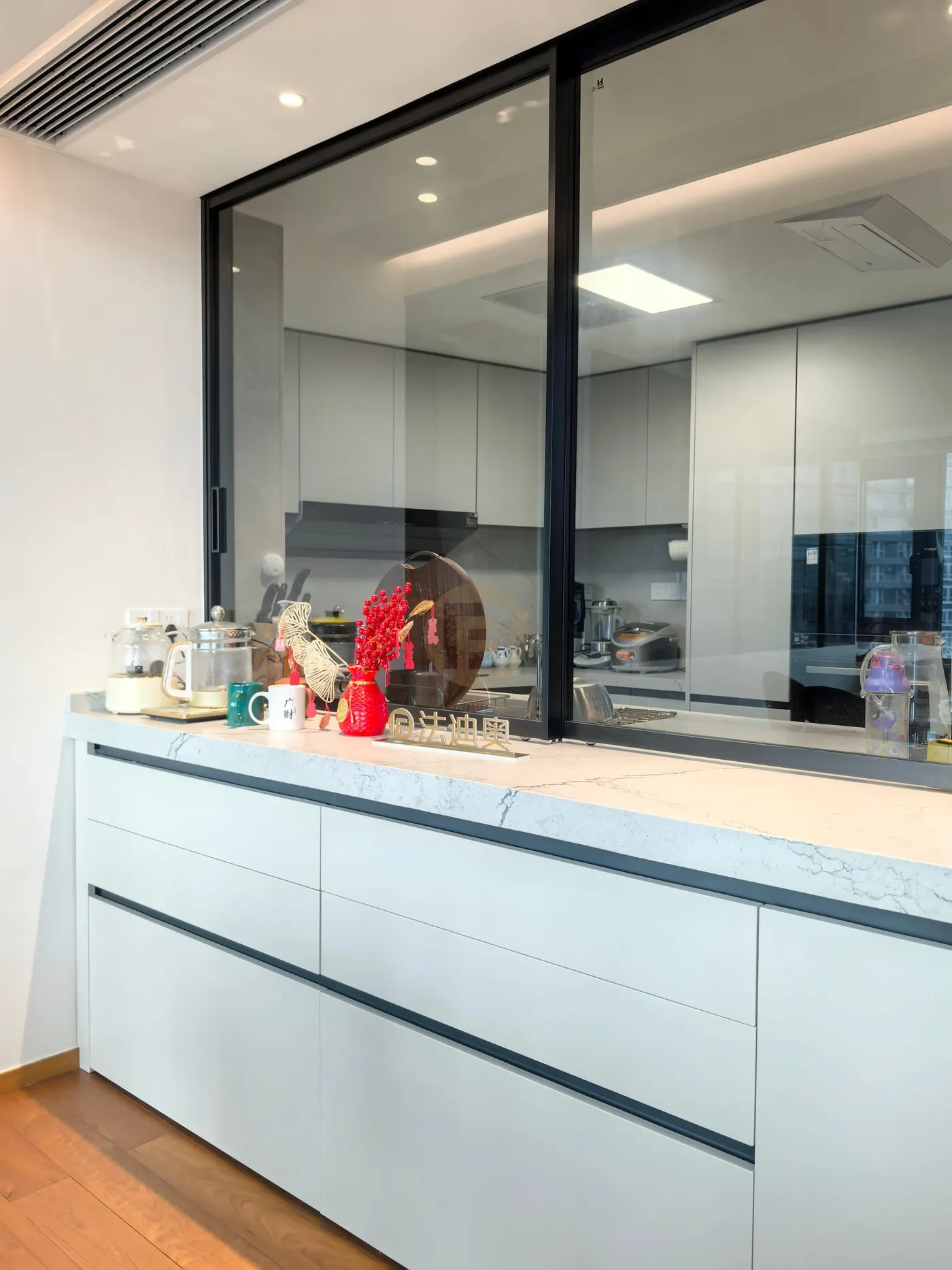 Stainless steel servery cabinetry with marble worktop below a black-framed pass-through window