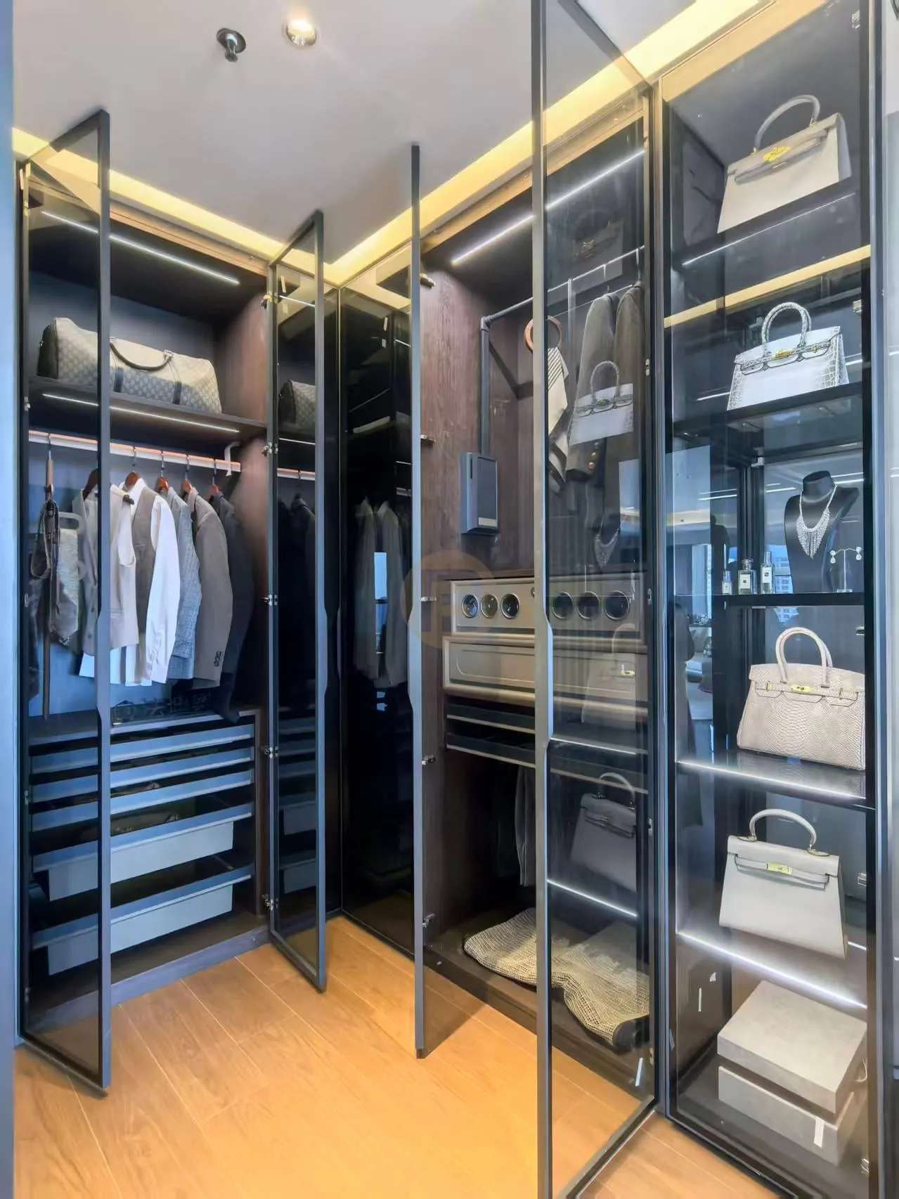 Glass-fronted walk-in closet with backlit shelving displaying handbags, watches and tailoring