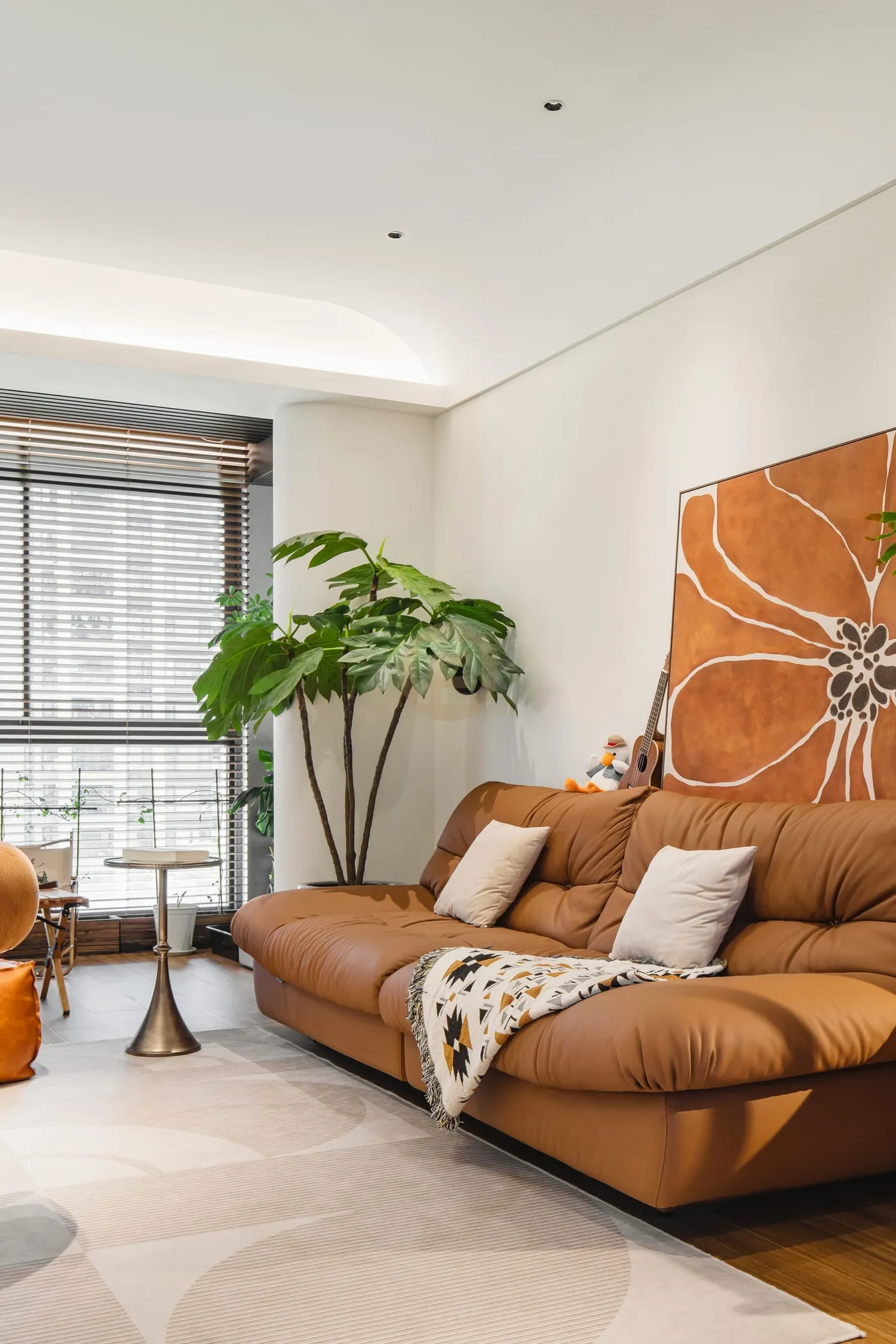Caramel leather sofa beneath large abstract art beside a plant-filled balcony with venetian blinds