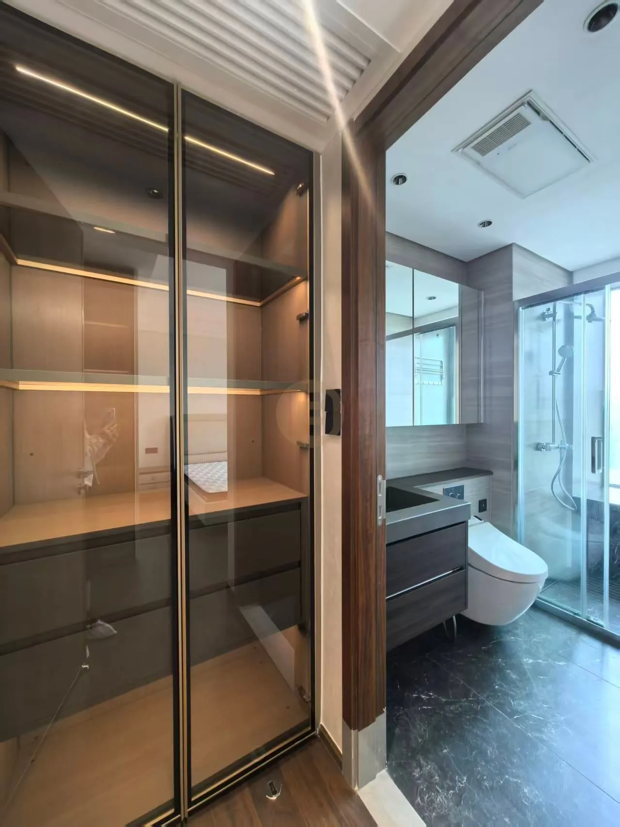 Glass-front linen cabinet beside a bathroom with dark wood-grain vanity, mirror wall and walk-in shower