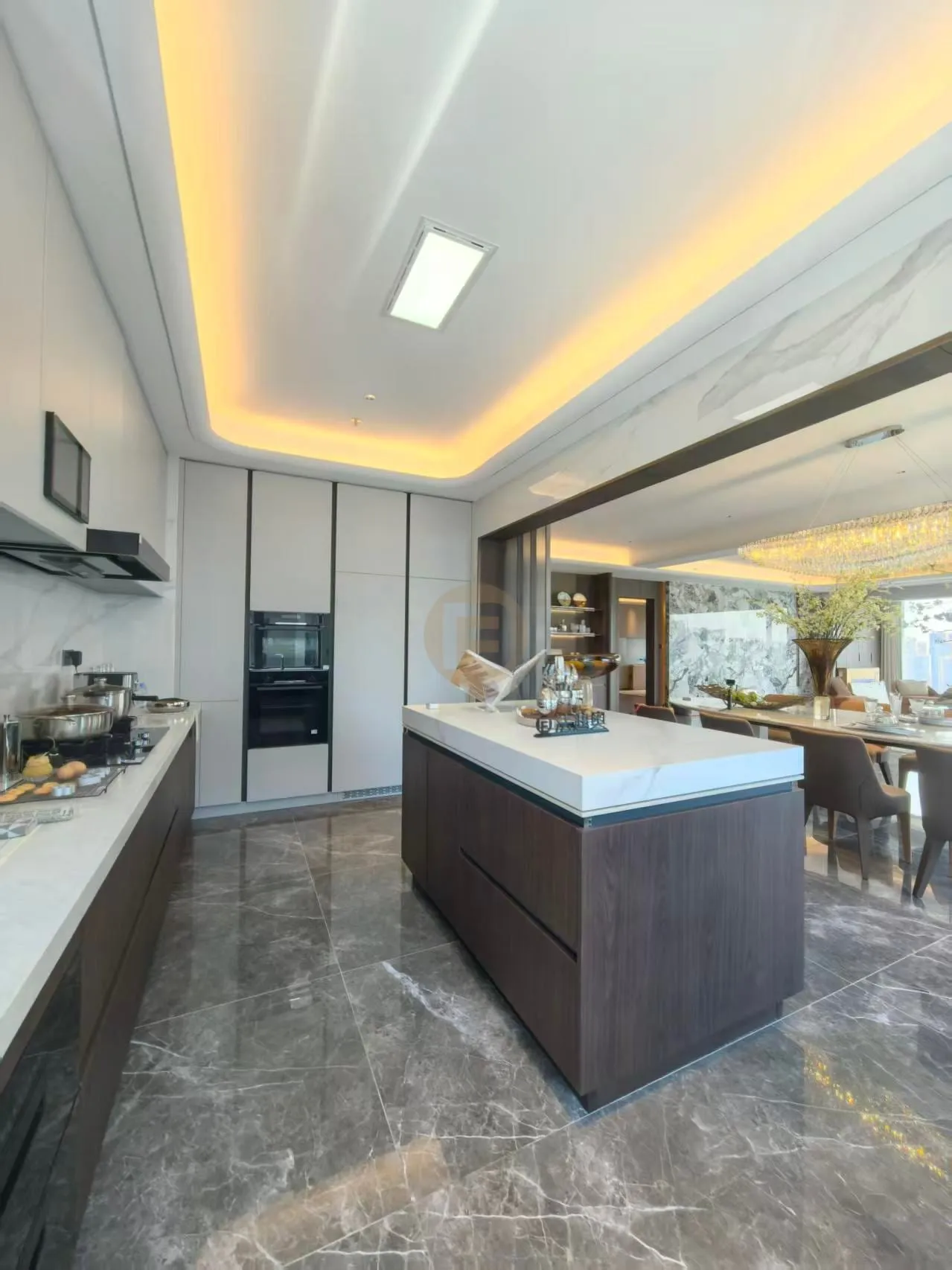 Island kitchen in walnut-tone stainless steel with white stone tops, open to a chandelier-lit dining room