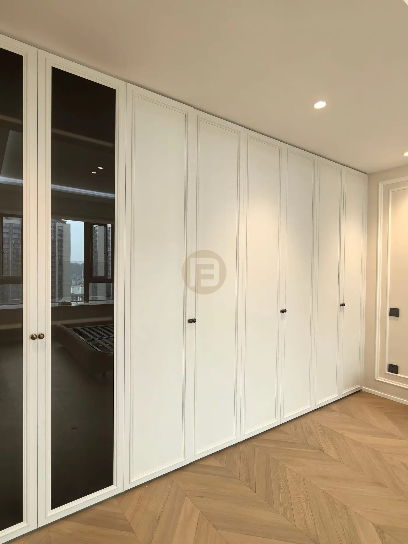 Floor-to-ceiling white paneled wardrobe wall with smoked glass doors over herringbone oak flooring