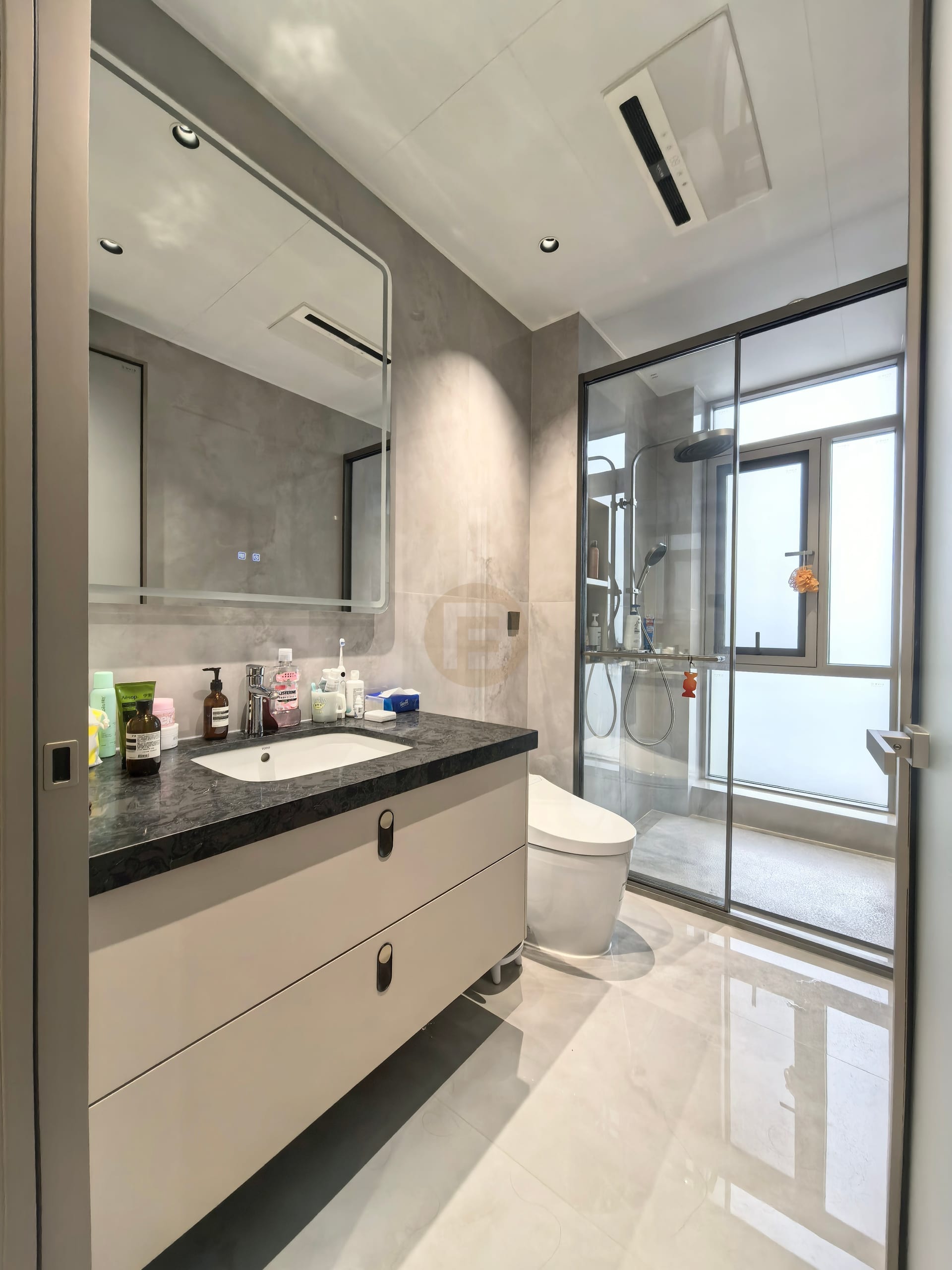 Wall-hung vanity with dark stone top and lit mirror beside a framed glass shower