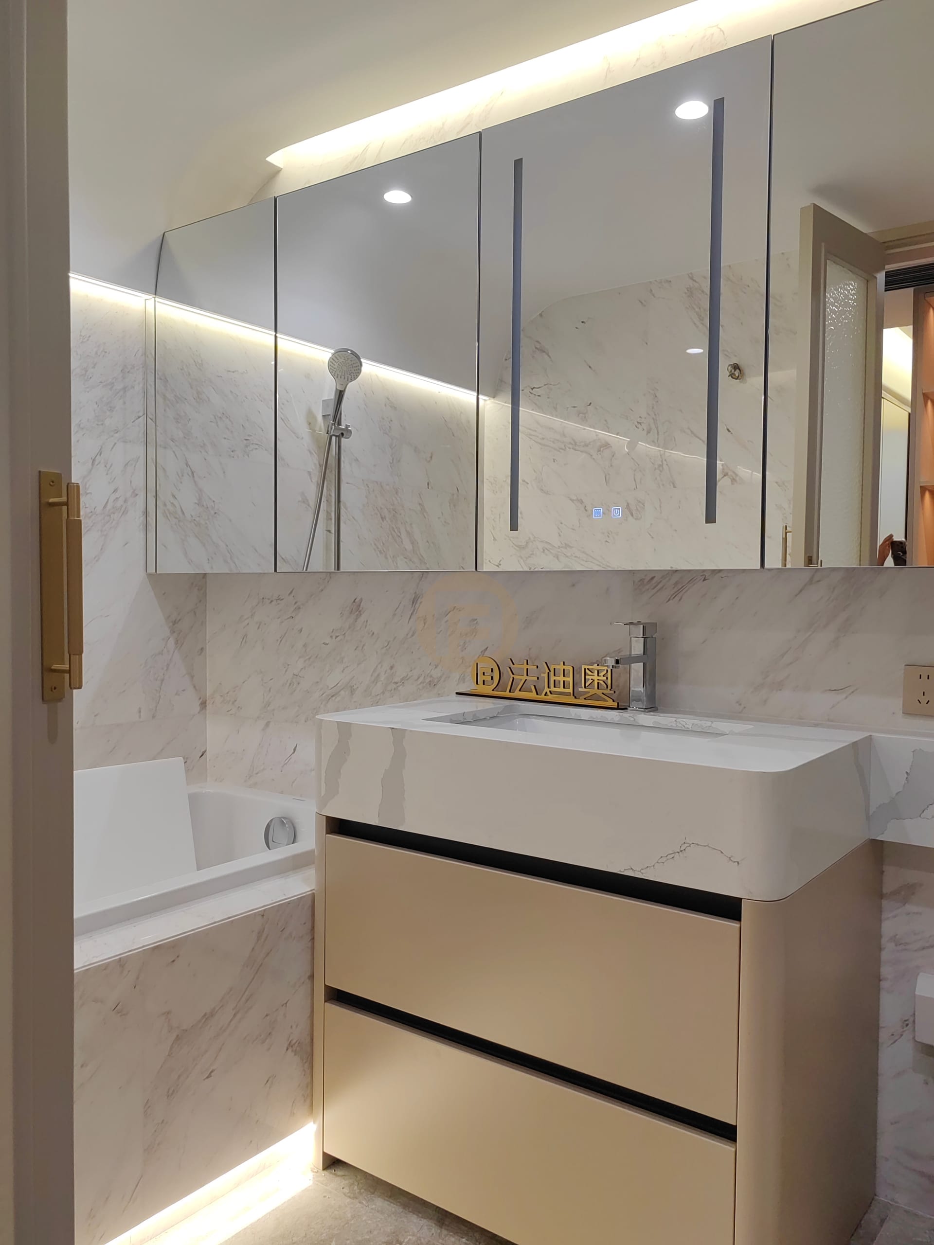 Cream stainless steel bathroom vanity with stone top, mirrored cabinet wall, brass pull and bathtub beyond