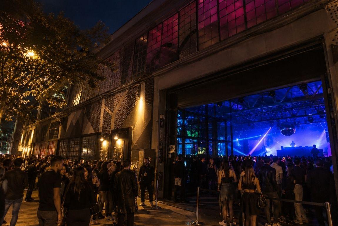 Crowds gathering outside a large nightclub at night, with blue and purple lights illuminating the dance floor inside.