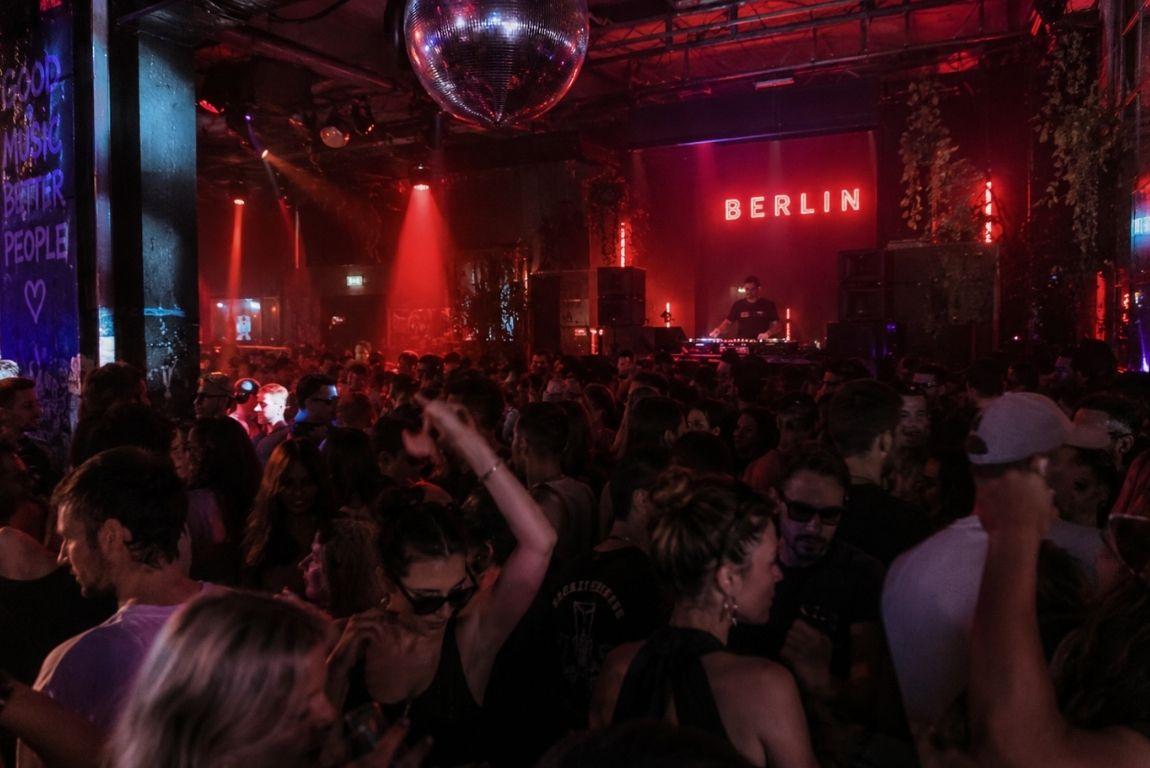 Crowded Berlin nightclub with people dancing under red lights while a DJ performs beneath a disco ball.