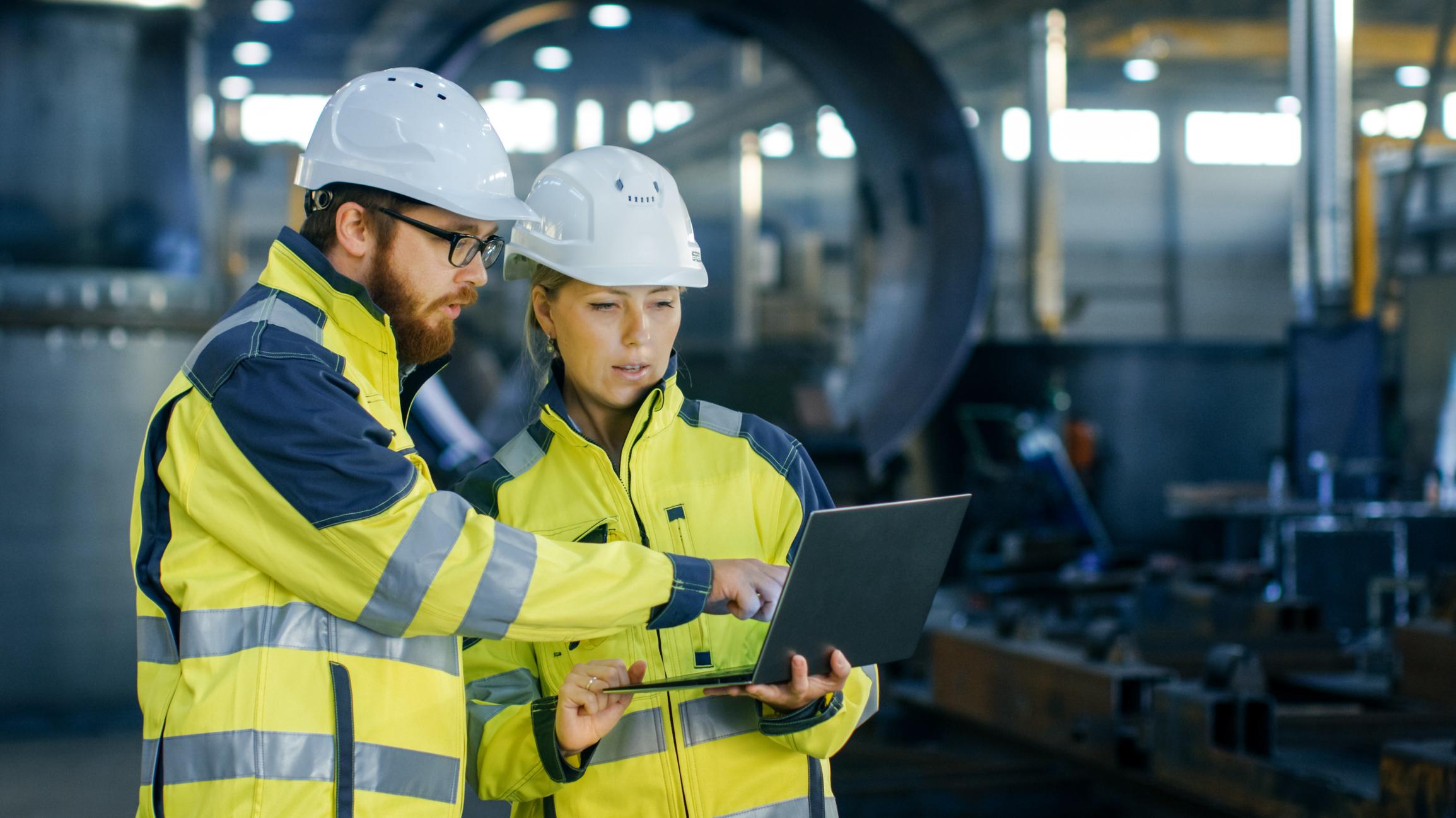 Two workers in hard hats and high-visibility jackets using a laptop beside machinery in an industrial facility.