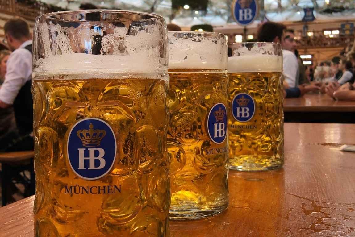 Three large Hofbräu beer steins on a table inside a crowded Oktoberfest beer tent in Munich.