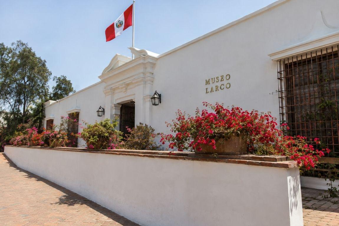 Exterior of Museo Larco in Lima, Peru, with white colonial architecture, colorful flowers, and the Peruvian flag.