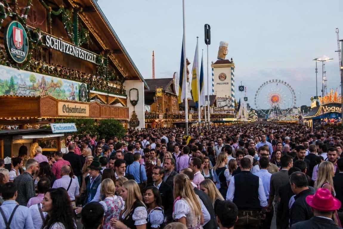 Large crowds walking through Oktoberfest in Munich, surrounded by beer tents, festival stalls, and a Ferris wheel.