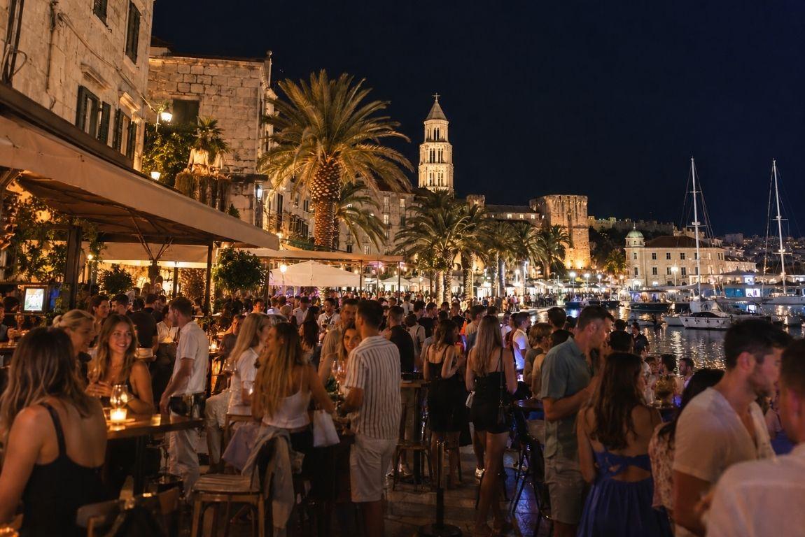Crowds enjoying nightlife at waterfront bars in Split, Croatia, with palm trees, yachts, and historic buildings illuminated at night.