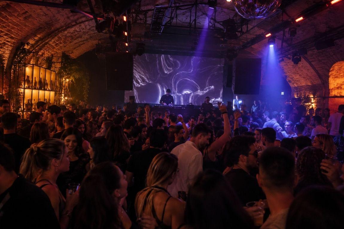 Crowded nightclub with people dancing under purple and red lights while a DJ performs beneath a disco ball.