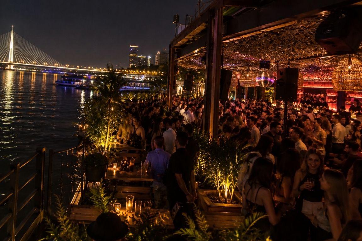 Crowds enjoying nightlife at a lively riverside bar, with a brightly lit cable-stayed bridge and city skyline in the background.