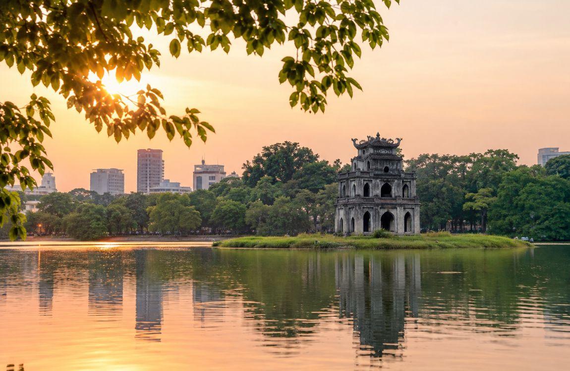 Turtle Tower on Hoan Kiem Lake in Hanoi at sunset, surrounded by trees with the city skyline in the background.