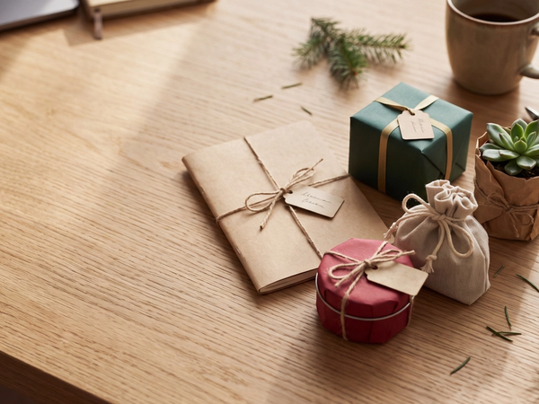 Cinq petits cadeaux emballés de papier kraft, vert et rouge, avec étiquettes vierges, sur un bureau en bois près d'un mug.