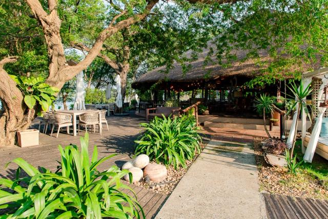 Restaurant deck with tables under the trees, leading to the thatched restaurant