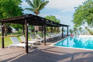 Swimming pool with sun loungers under a wooden pergola, palm trees and the lagoon beyond