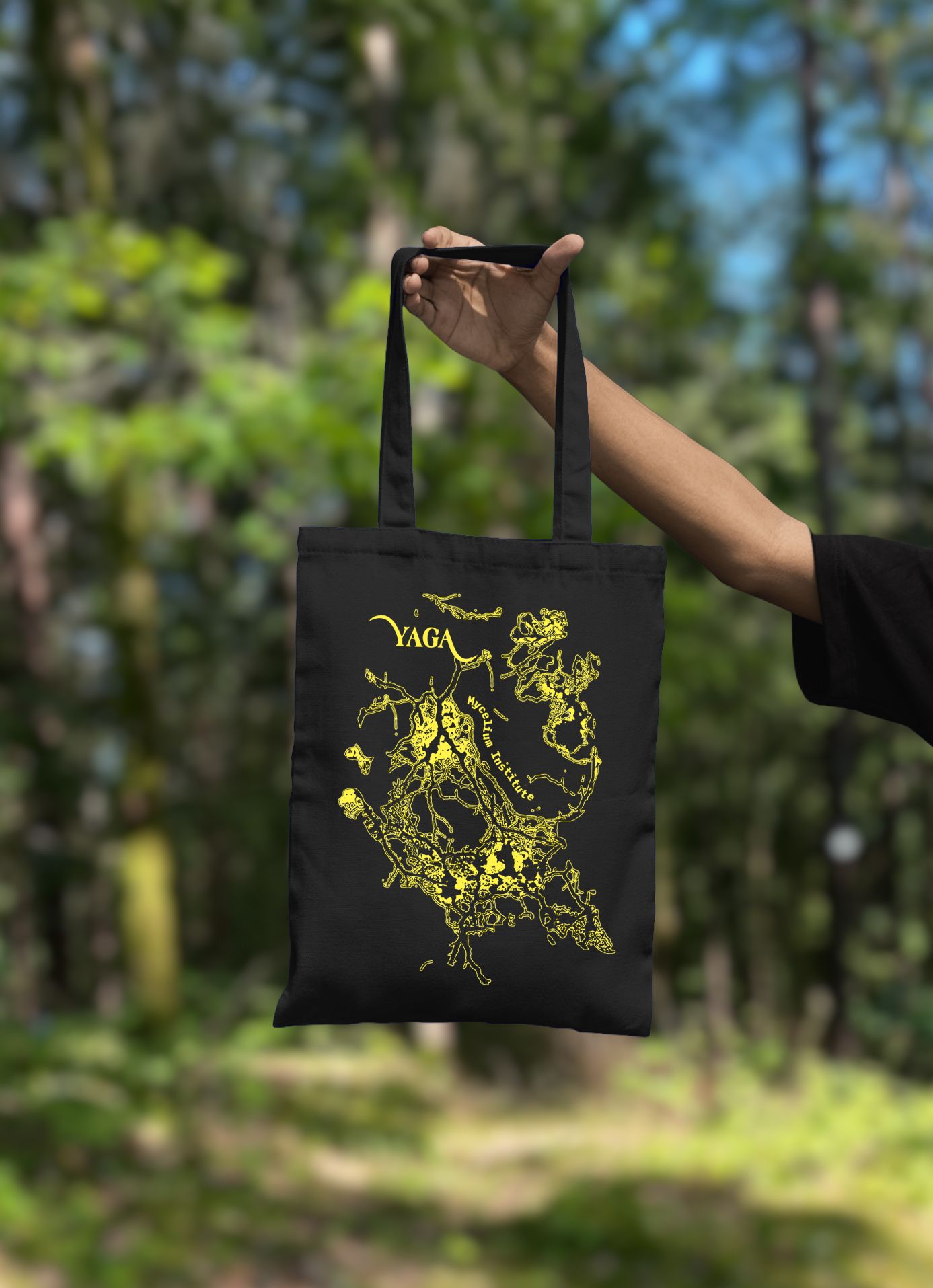 Black YAGA tote bag with a yellow mycelium print held up in a forest
