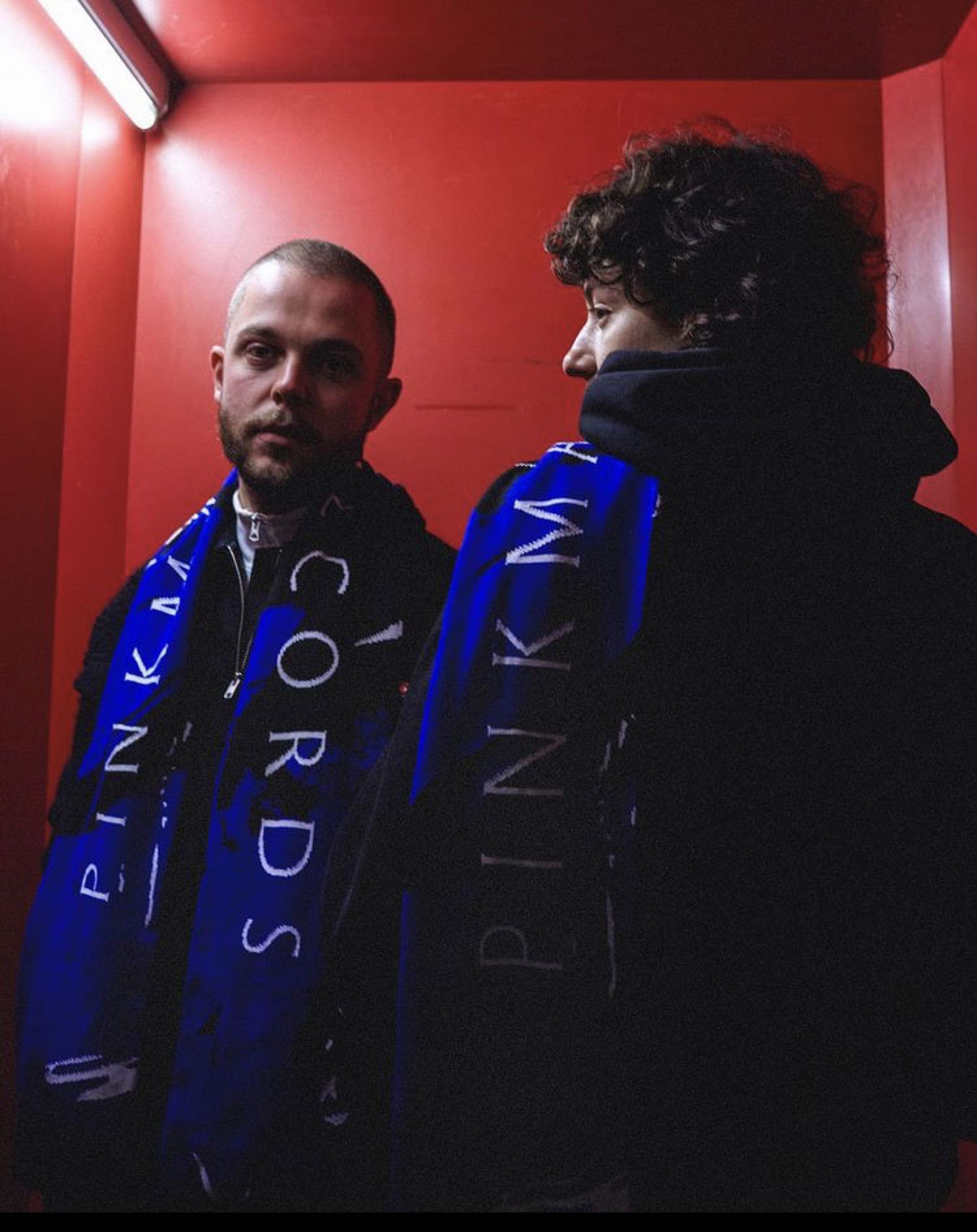 Two people wearing blue Pinkman Records scarves in a red room