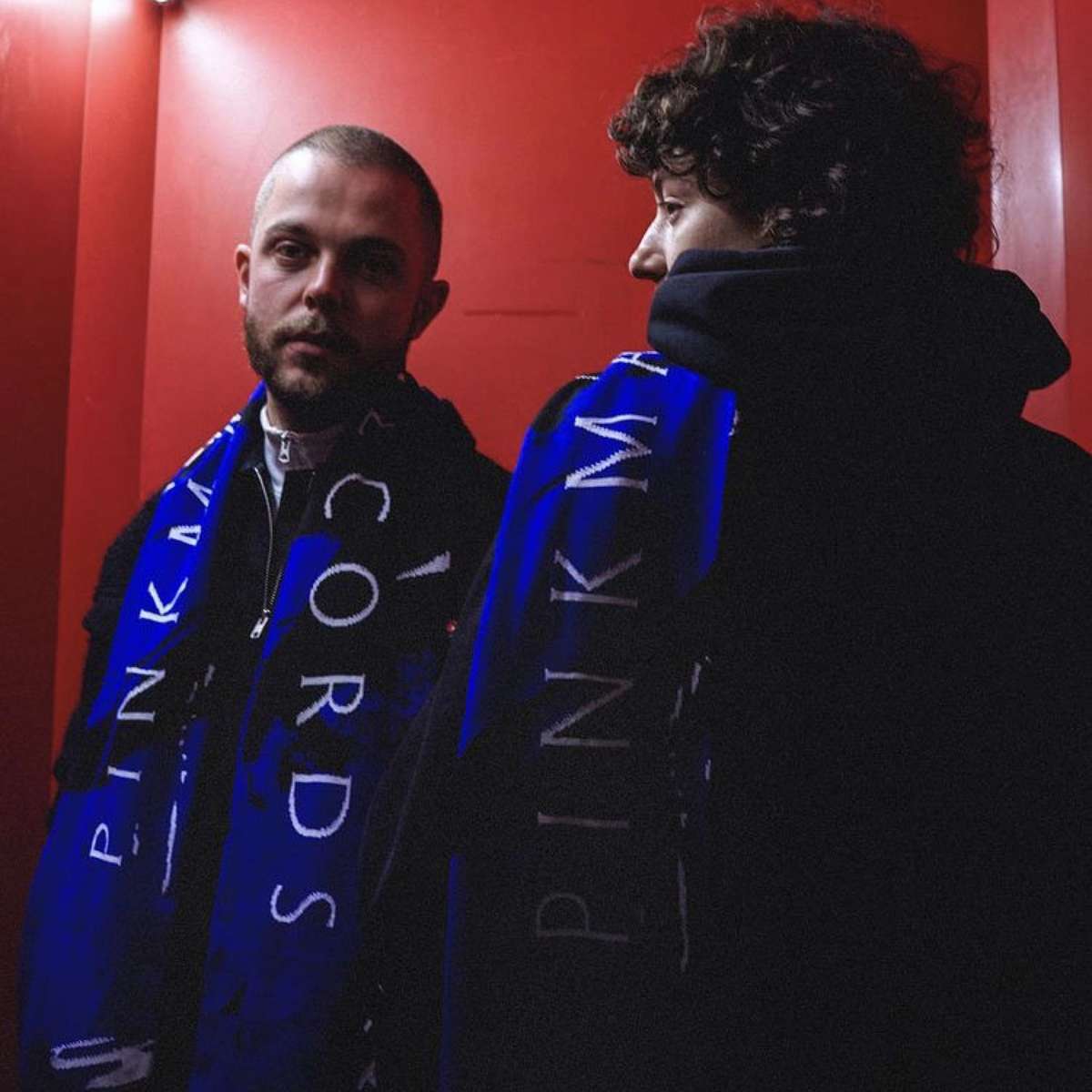 Two people wearing blue Pinkman Records scarves in a red room