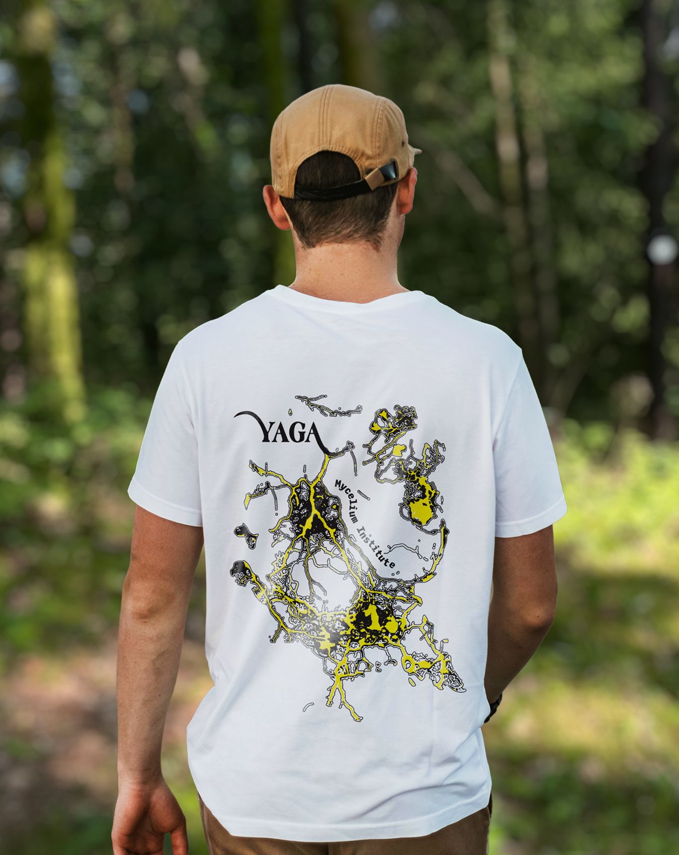 A person in a white YAGA T-shirt with a mycelium print on the back, seen from behind in a forest