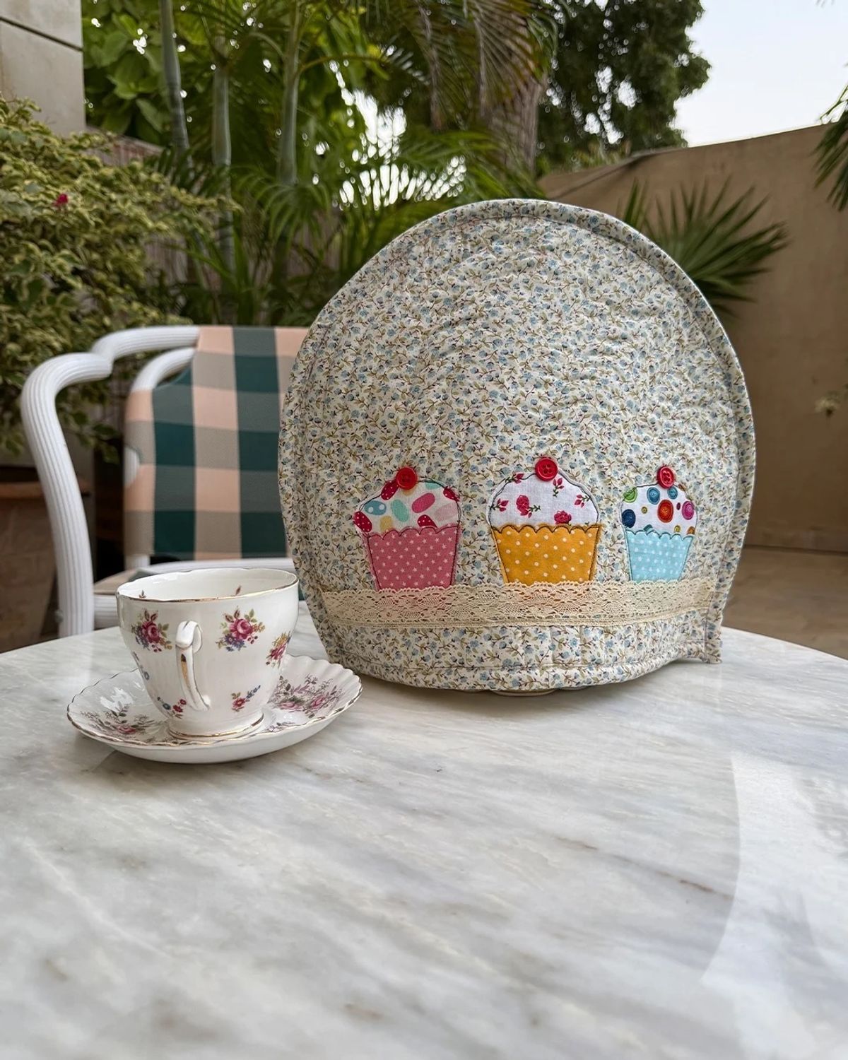 Quilted tea cosy with three appliqué cupcakes beside a china teacup