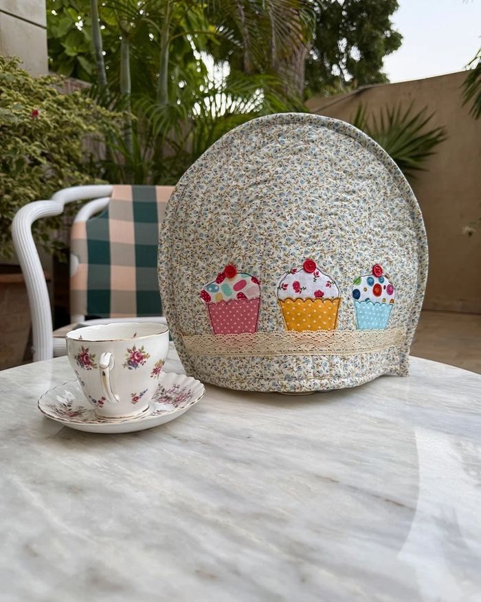 Quilted tea cosy with three appliqué cupcakes beside a china teacup