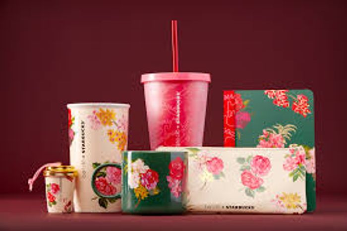  A visually appealing collection of Starbucks merchandise against a dark red background. The items are primarily decorated with floral patterns featuring red, pink, and yellow flowers on white and dark green bases. The collection includes a tall white tumbler, a tall pink cold cup with a straw, a small gold and white keychain cup, a dark green mug, a floral clutch or pouch, and a matching floral notebook or journal. The image promotes the brand's seasonal or special edition accessories and drinkware.