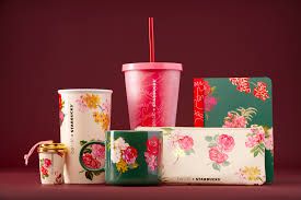 A visually appealing collection of Starbucks merchandise against a dark red background. The items are primarily decorated with floral patterns featuring red, pink, and yellow flowers on white and dark green bases. The collection includes a tall white tumbler, a tall pink cold cup with a straw, a small gold and white keychain cup, a dark green mug, a floral clutch or pouch, and a matching floral notebook or journal. The image promotes the brand's seasonal or special edition accessories and drinkware.