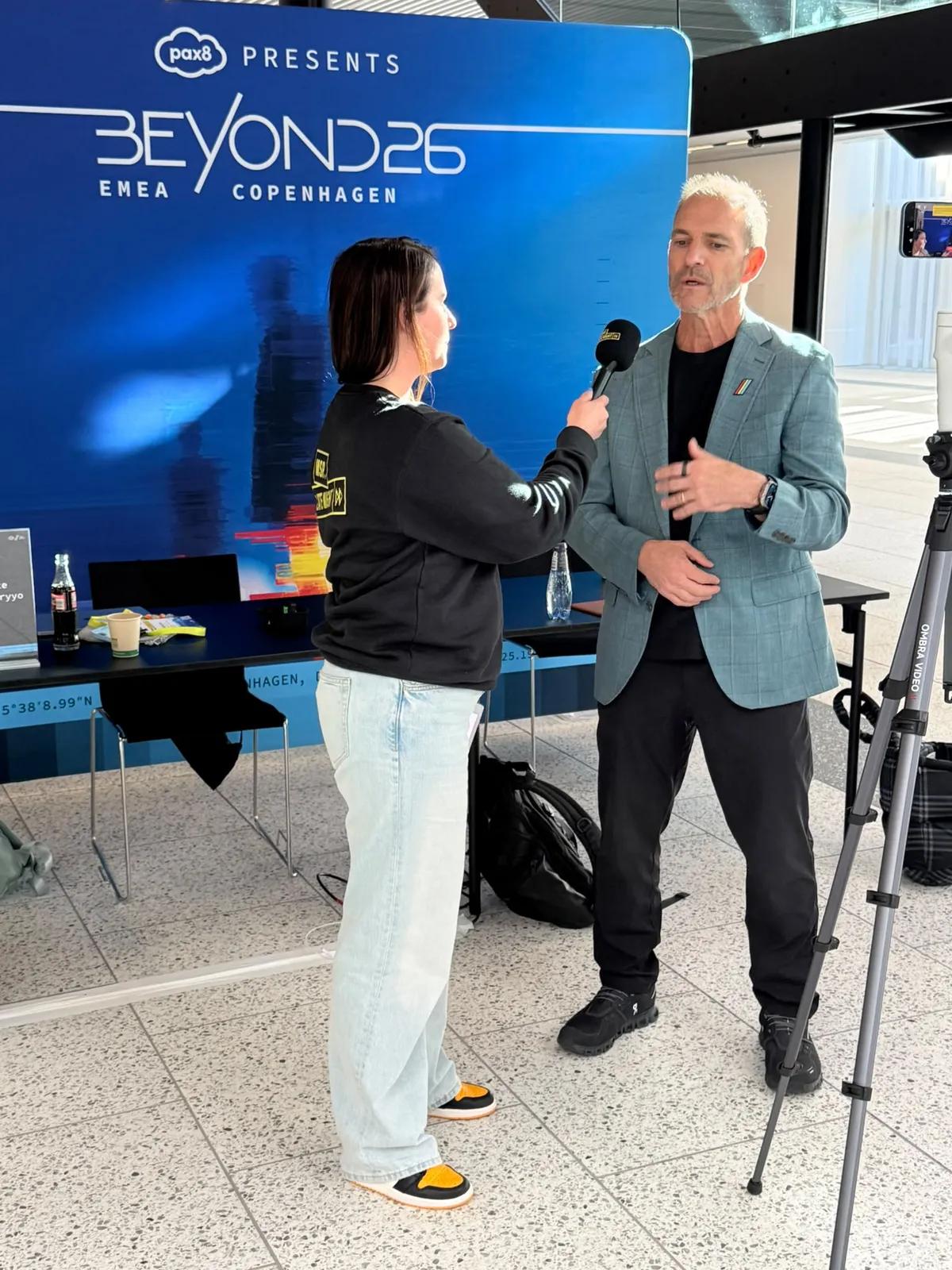 MSP Late Night interviews a guest with microphone and camera in front of the Pax8 Beyond26 wall in Copenhagen