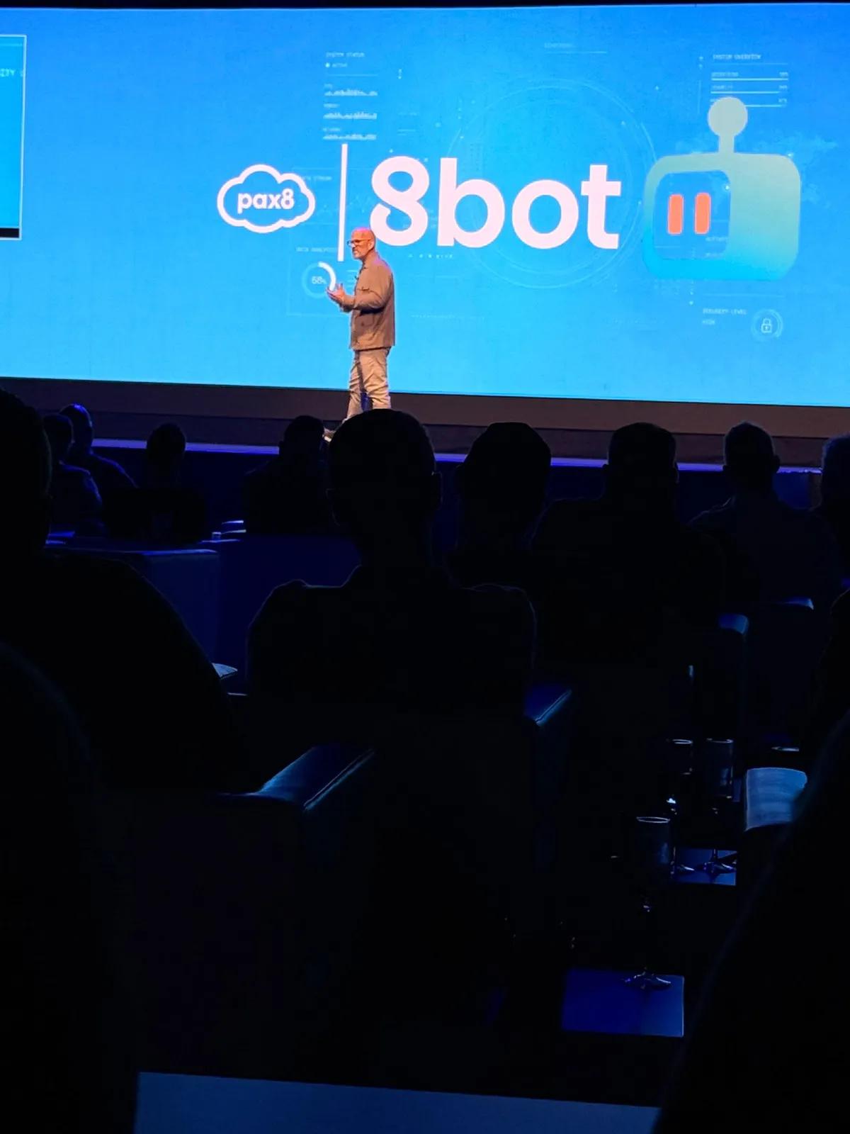 A speaker on the Pax8 Beyond EMEA stage in front of a blue screen with Pax8 8bot