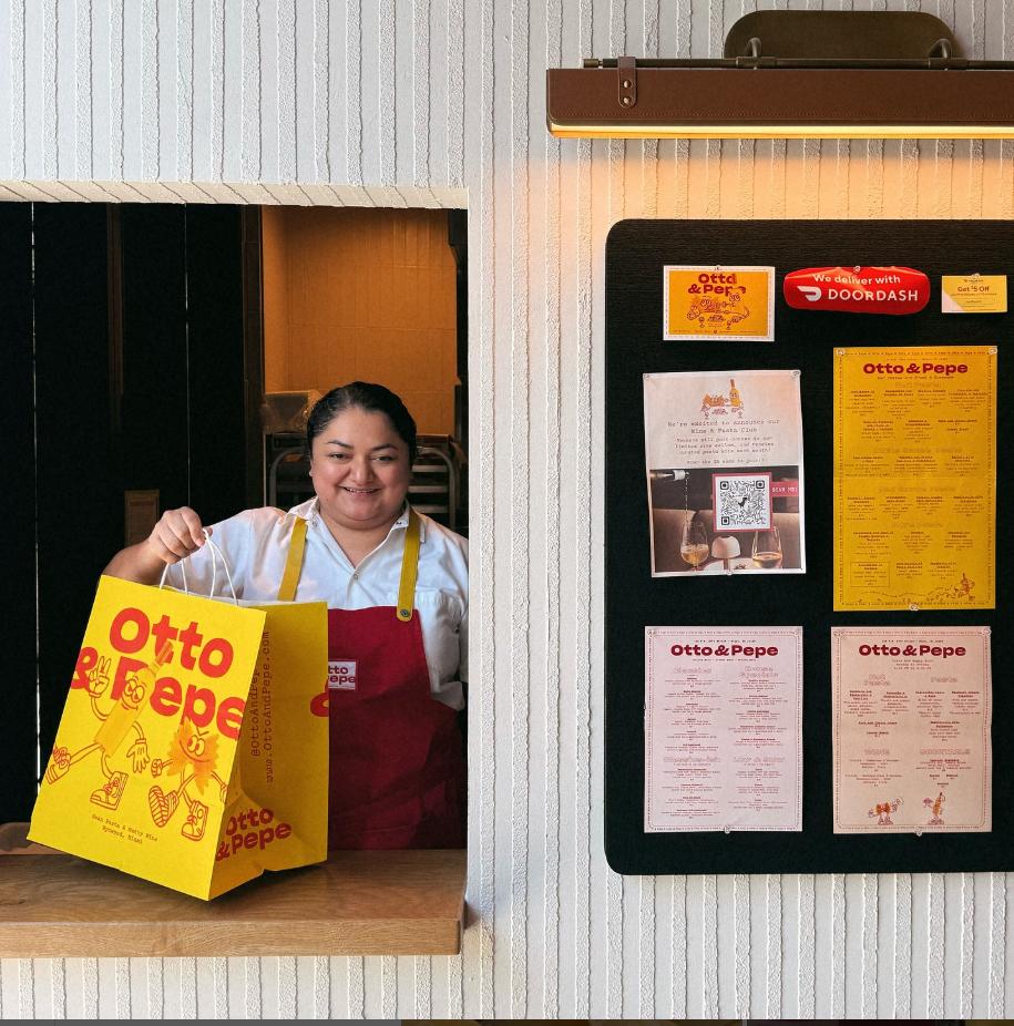 An Otto & Pepe staff member presents branded takeout packaging from the service window inside the Wynwood, Miami pasta bar and natural wine shop. The bright yellow packaging, illustrated characters, menus, and printed collateral showcase the playful visual identity developed by Love & War, extending the Otto & Pepe brand across every part of the restaurant experience.