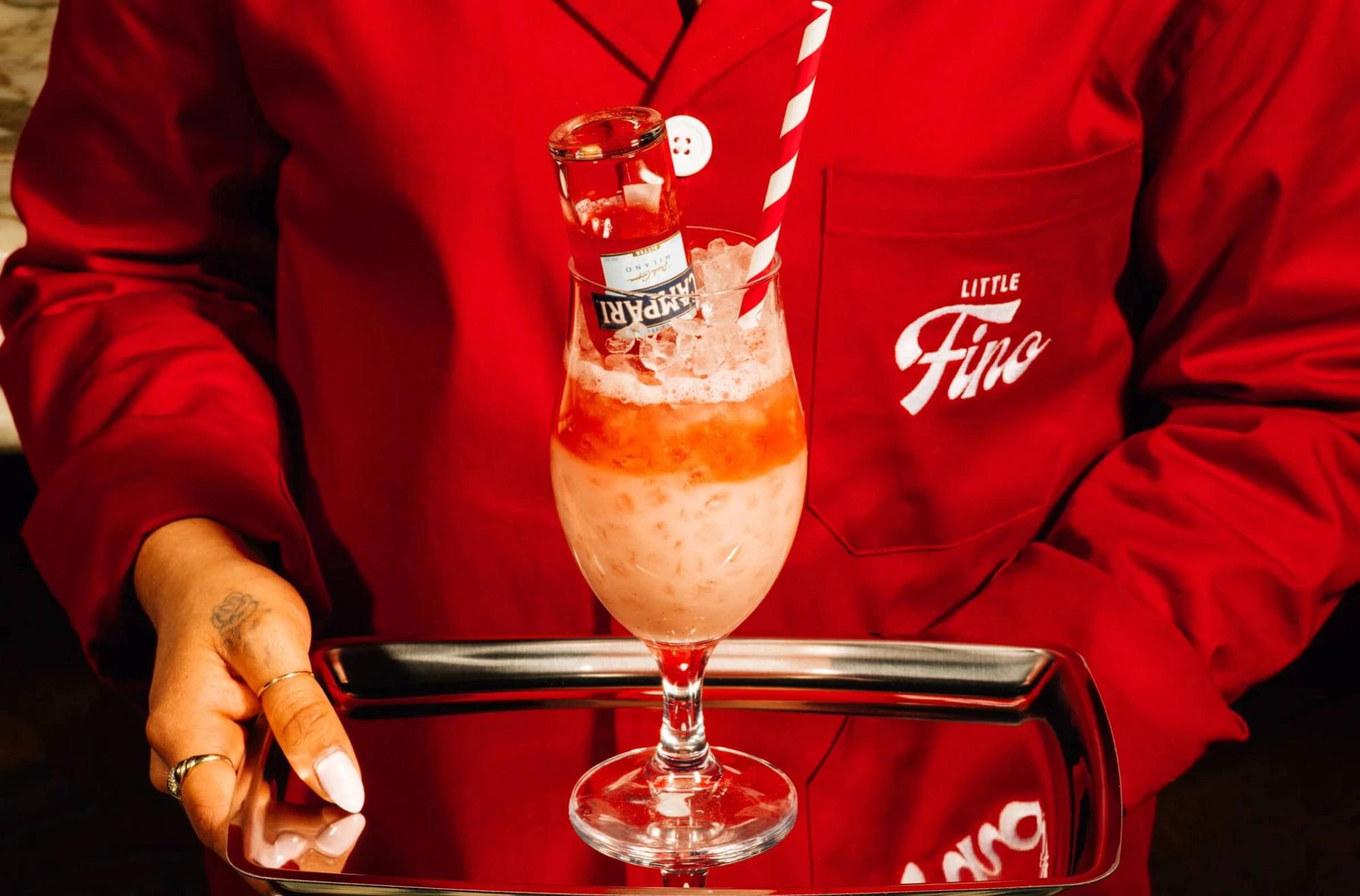 Signature cocktail at Little Fino, the all-day Italian café and bar at The William Vale in Williamsburg, Brooklyn, served by staff in a custom red Little Fino uniform with branded tray and striped straw. Love & War developed the name, brand concept, visual identity and branded experience for Little Fino, extending the identity across uniforms, cocktails, menus, packaging, signage, serveware and custom illustrations to create a distinctive contemporary take on Italian café and bar culture.