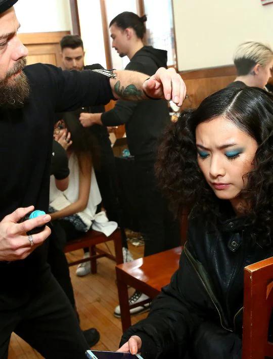 Stylist John D applying product to a model's hair, with other models and stylists in the background