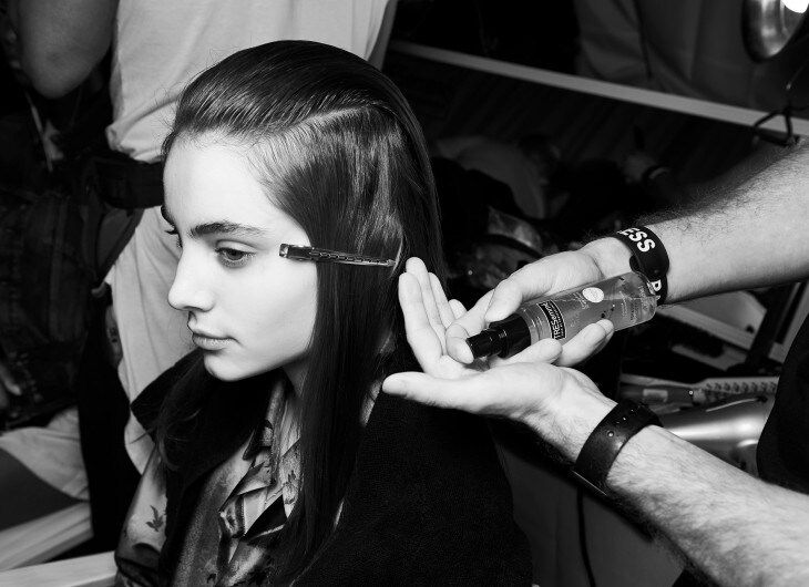 A model with her hair held in place with a clip, and a stylist applying product to it