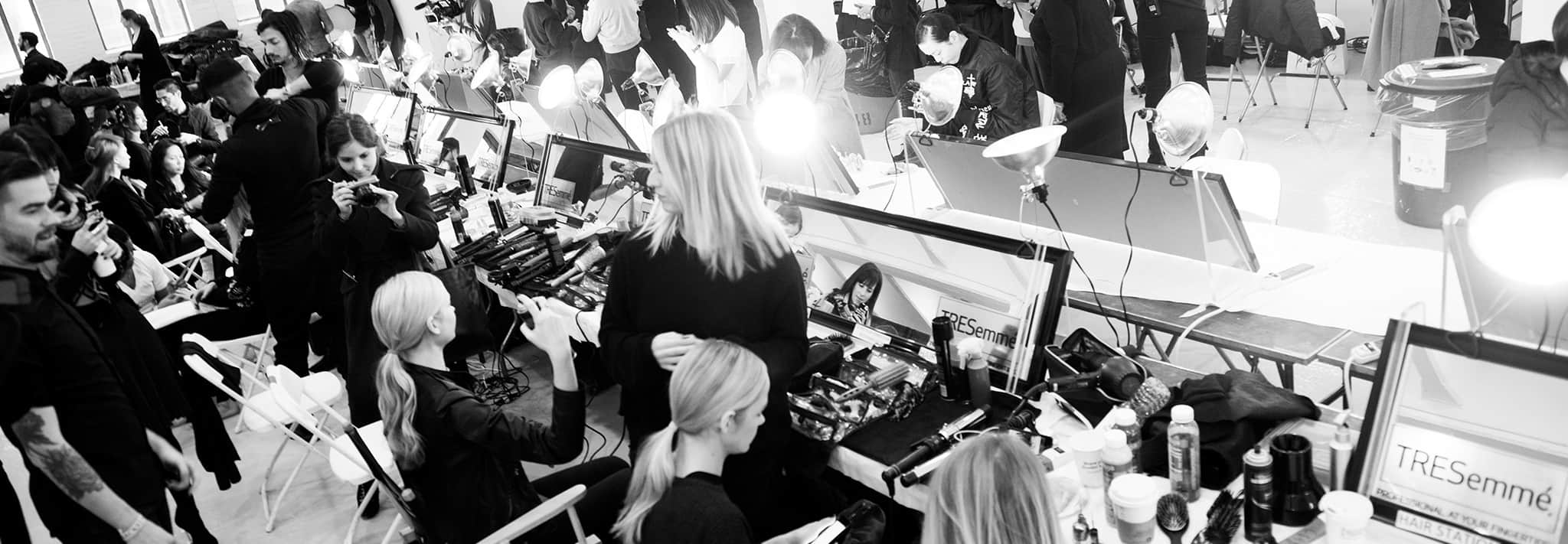 Black and white shot of a busy backstage area featuring models sitting in front of mirrors with hairstylists, photographers and styling tools and hair products