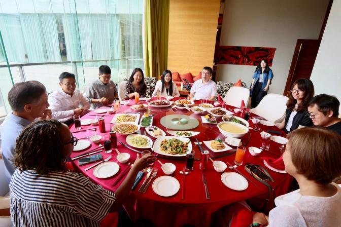 Group of people gathered around a large round red table with a lazy susan, sharing Chinese dishes in a modern dining room.
