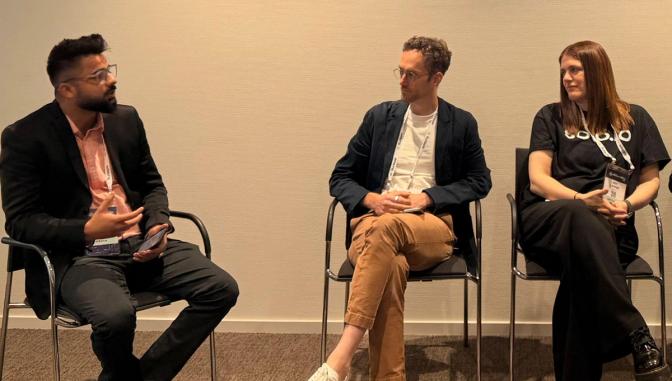 Three panelists seated on chairs at a conference, with the man on the left speaking while the other two listen.