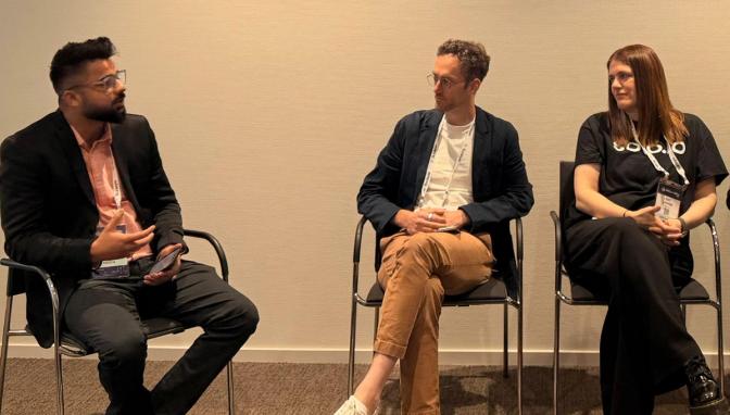 Three panelists seated on chairs at a conference, with the man on the left speaking while the other two listen.