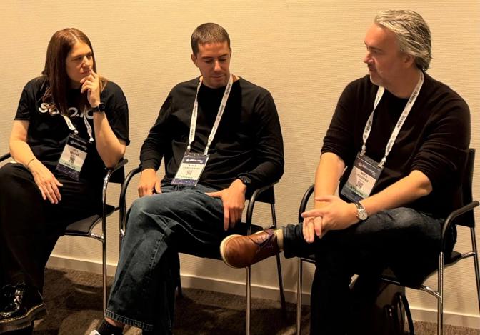 Three conference attendees wearing lanyards and black tops sit in chairs having a discussion at a tech event.