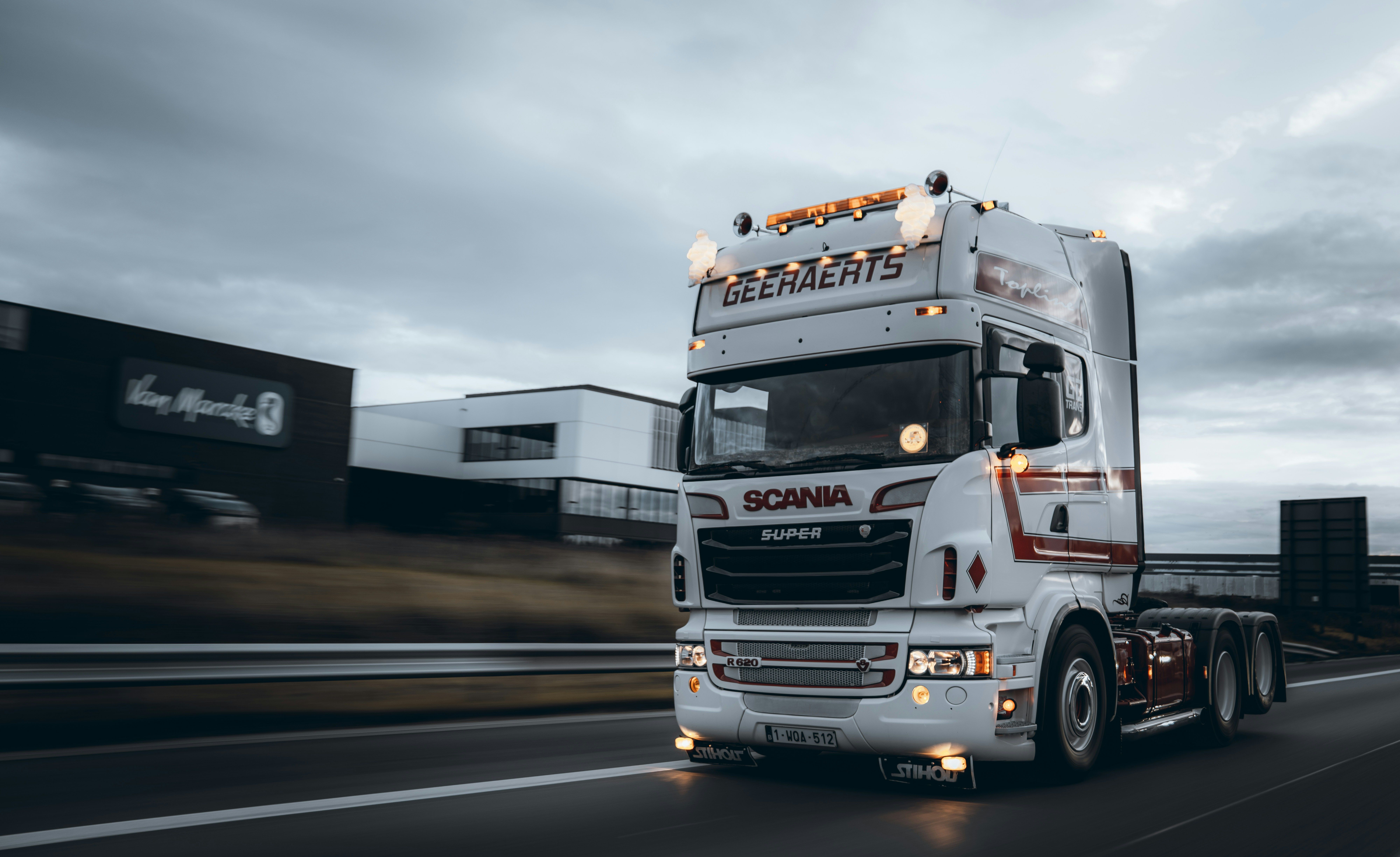 Biała ciężarówka Scania Super R620 z napisem „GEERAERTS” na daszku przeciwsłonecznym i włączonymi światłami, pędząca lewym pasem autostrady, z rozmytym tłem budynków przemysłowych i barierą energochłonną pod zachmurzonym niebem.