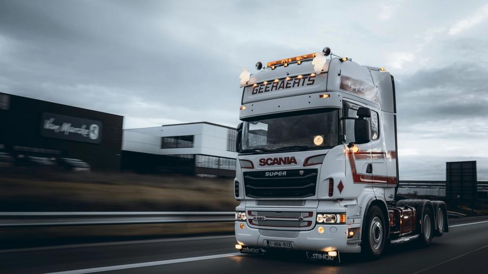 Biała ciężarówka Scania Super R620 z napisem „GEERAERTS” na daszku przeciwsłonecznym i włączonymi światłami, pędząca lewym pasem autostrady, z rozmytym tłem budynków przemysłowych i barierą energochłonną pod zachmurzonym niebem. Biała ciężarówka Scania Super R620 z napisem „GEERAERTS” na daszku przeciwsłonecznym i włączonymi światłami, pędząca lewym pasem autostrady, z rozmytym tłem budynków przemysłowych i barierą energochłonną pod zachmurzonym niebem.