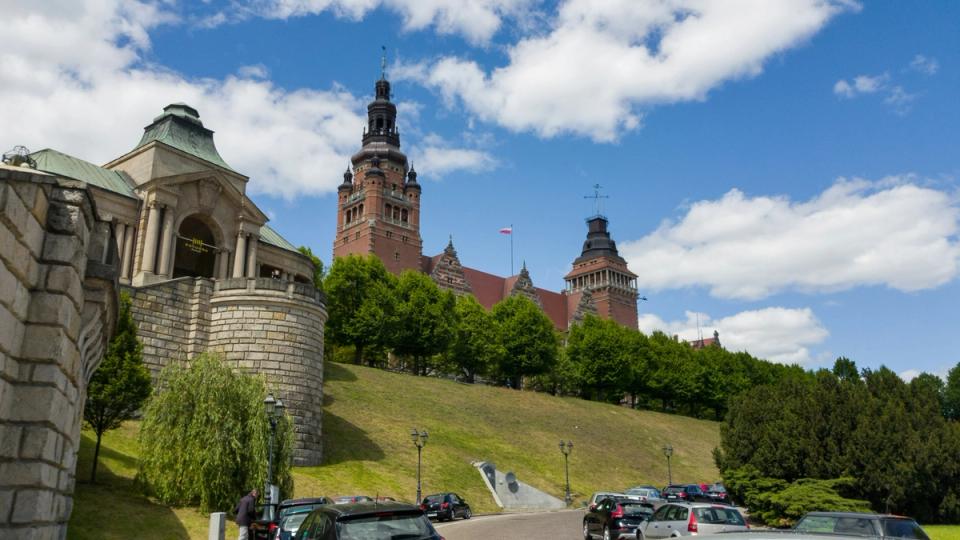 Gmach Urzędu Wojewódzkiego i Muzeum Narodowego na Wałach Chrobrego w Szczecinie, widziany z dołu, z zabytkową architekturą, wieżami i otaczającą zielenią, na tle nieba z białymi chmurami.