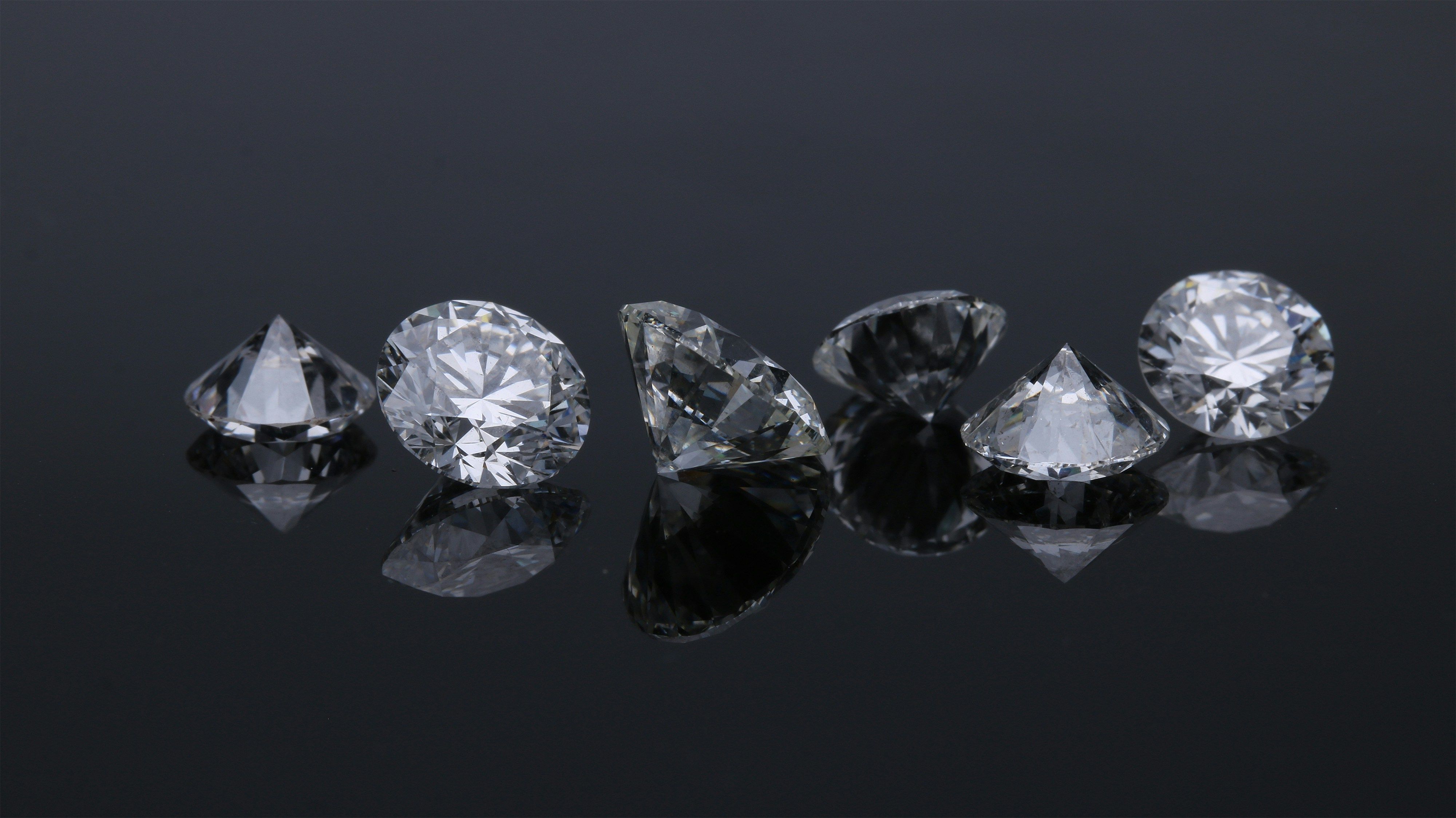 Fotografia produktowa przedstawiająca sześć luksusowych, precyzyjnie oszlifowanych diamentów o brylantowym szlifie, ułożonych w rzędzie na czarnym, lustrzanym tle. Kamienie szlachetne mocno błyszczą, odbijając światło w widoczny sposób, a ich perfekcyjne odbicia są widoczne na gładkiej powierzchni, co podkreśla ich wysoką wartość (High-Value).
