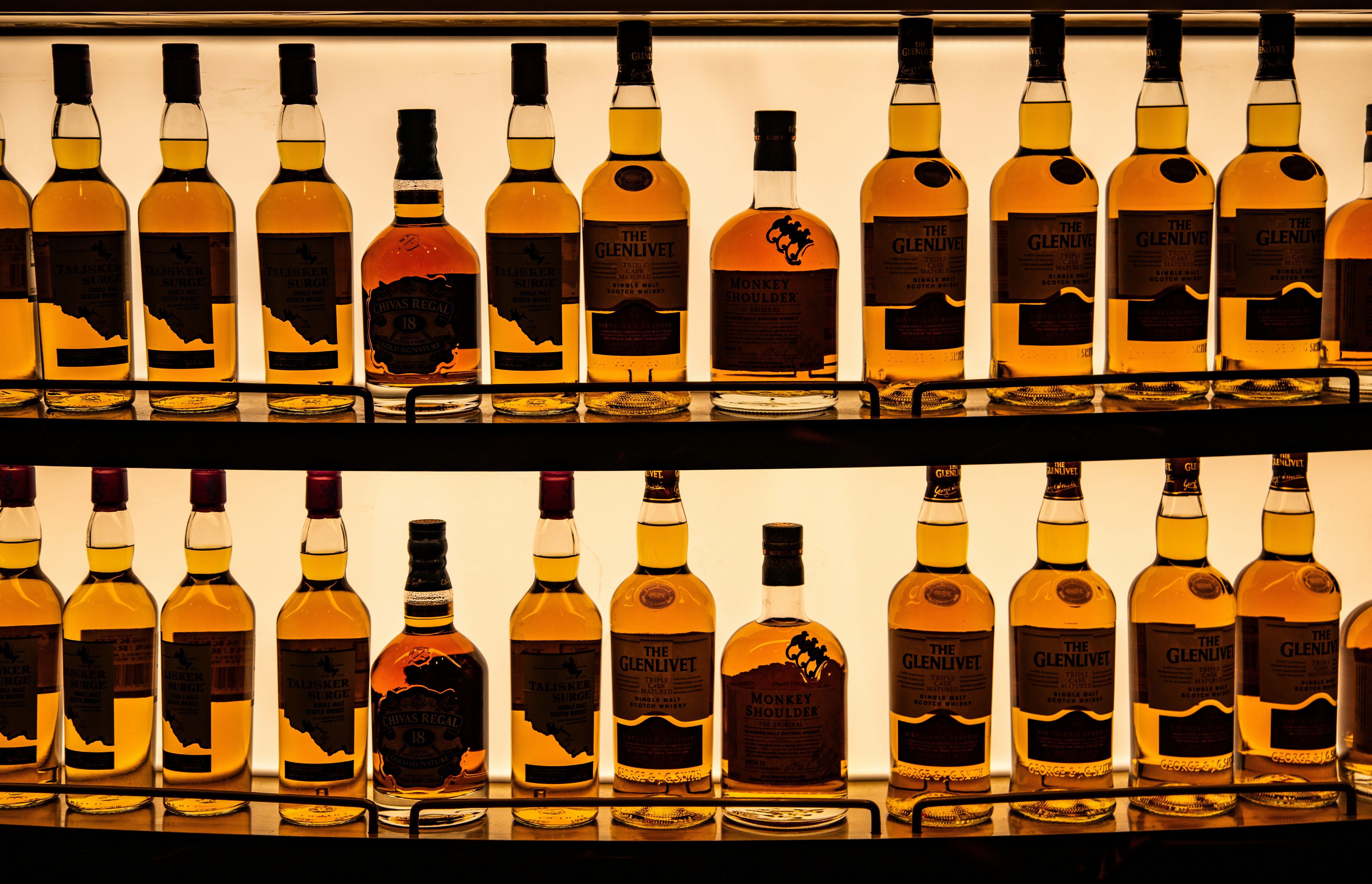 Podświetlone szklane półki wypełnione rzędami butelek whisky i ginu różnych marek w eleganckim barze. Idealne jako ilustracja asortymentu w logistyce napojów alkoholowych.