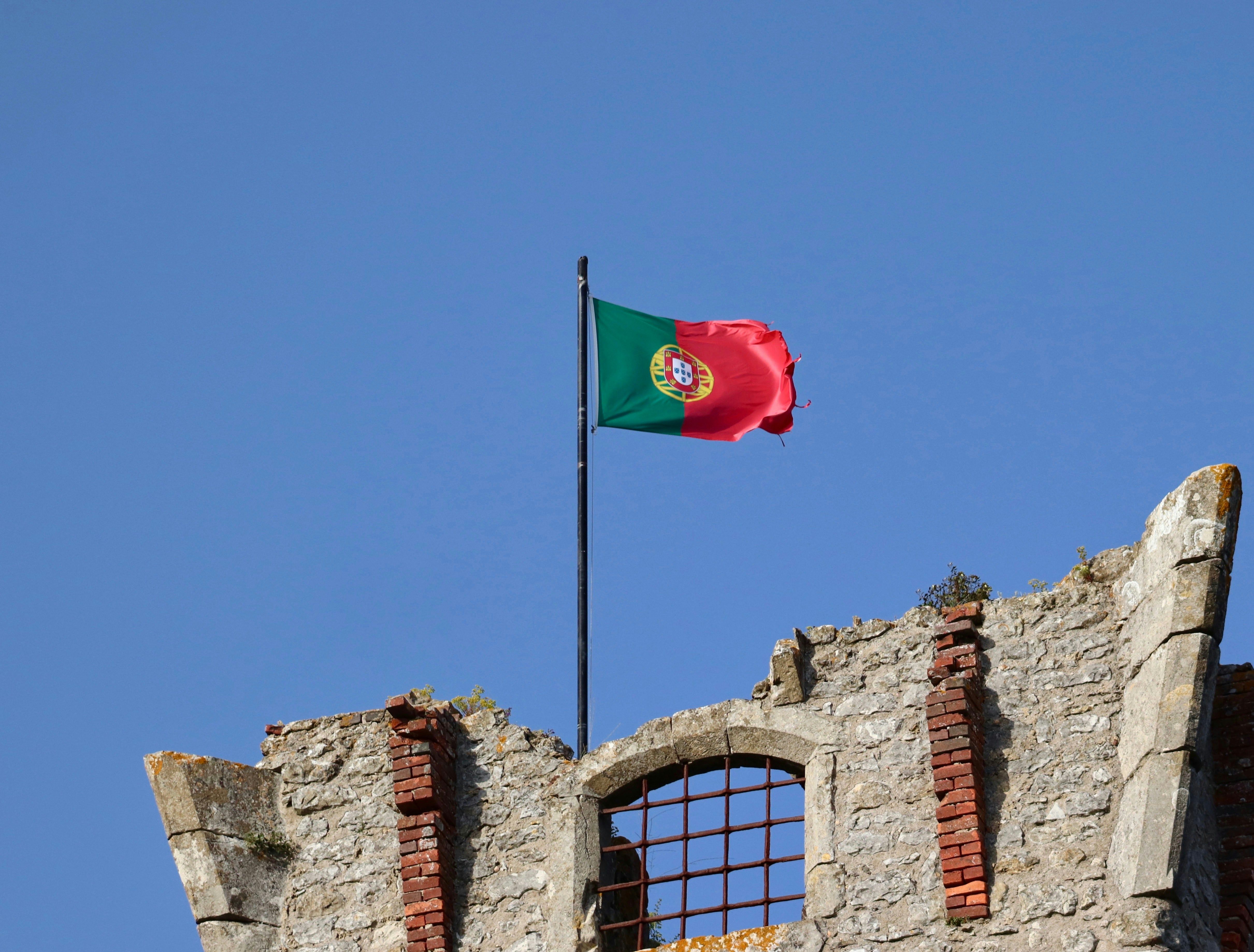 Przymocowana do nadgryzionego zębem czasu kamiennego fragmentu budynku flaga Portugalii powiewa na wietrze na tle niebieskiego, bezchmurnego nieba.