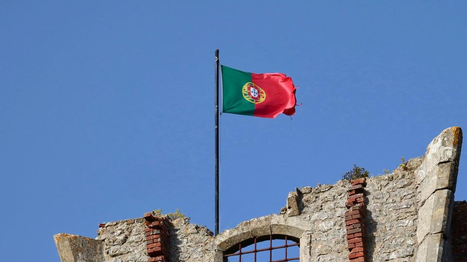Przymocowana do nadgryzionego zębem czasu kamiennego fragmentu budynku flaga Portugalii powiewa na wietrze na tle niebieskiego, bezchmurnego nieba. Przymocowana do nadgryzionego zębem czasu kamiennego fragmentu budynku flaga Portugalii powiewa na wietrze na tle niebieskiego, bezchmurnego nieba.
